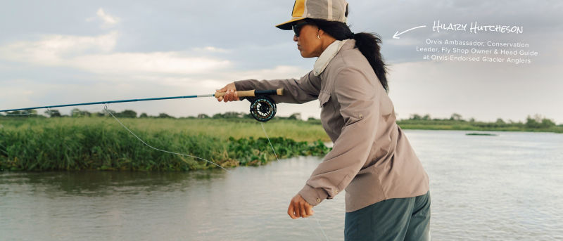 Am angler leans into her cast from the prow of a small boat on a placid river.