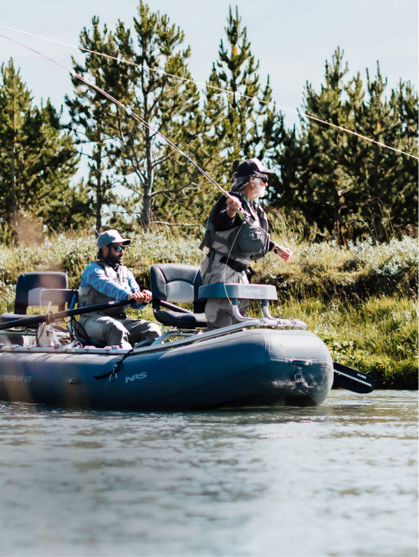 Two people fishing from an inflatable boat on a calm river surrounded by lush greenery and pine trees.