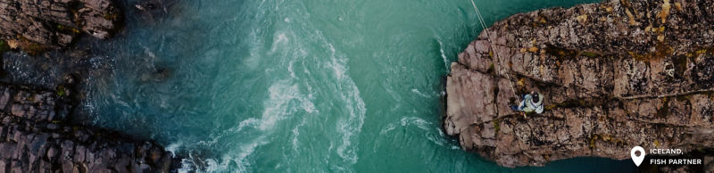 A stunning aerial view of a turquoise river winding through rugged rocky cliffs. An angler is standing on a prominent rock formation.