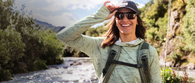 A smiling angler adjusts her ball cap in scenic river landscape.