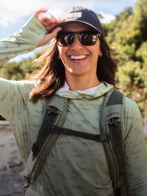 A smiling angler adjusts her ball cap in scenic river landscape.
