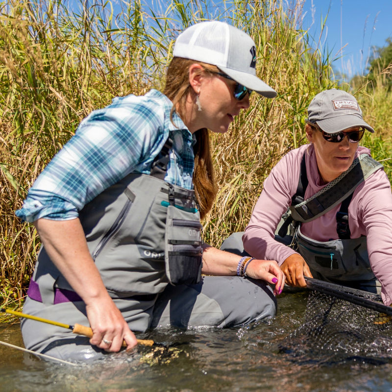 Two female anglers land a catch in a reedy river.