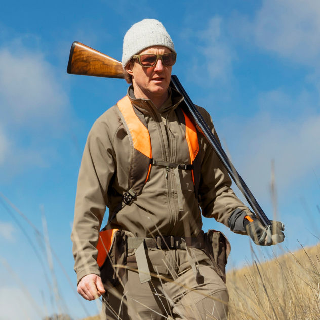 A hunter walks through dry grass under a bright blue sky.