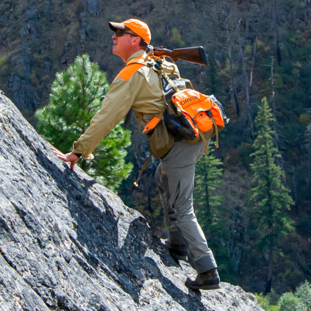 A hunter boulders up a steep rock incline holding his shotgun over his shoulder.