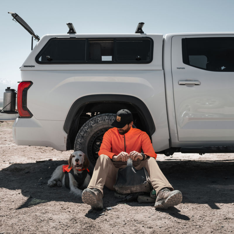 A hunter sits in the shade of his pickup truck with his bird dog.