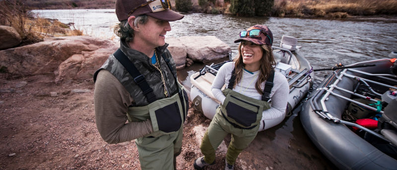 Two anglers dressed in fishing waders and outdoor gear, standing beside an inflatable raft on a riverbank.