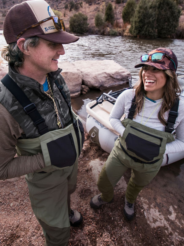 Two anglers dressed in fishing waders and outdoor gear, standing beside an inflatable raft on a riverbank.