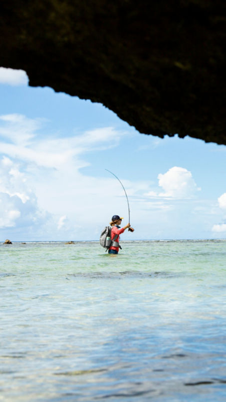 A view from under a rocky overhang of an angler casting her fly from hip-deep in the ocean.