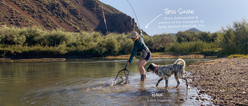 Jess Suvak stands in a shallow river netting a fish while her dog walks beside her in the water.