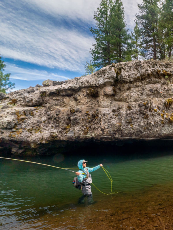 An angler casting from hip deep in a river wearing a sun protective shirt and waders.