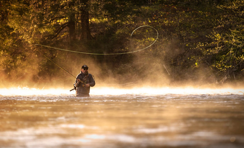 An angler casts his rod from hip-deep in a river with mist rising around him.