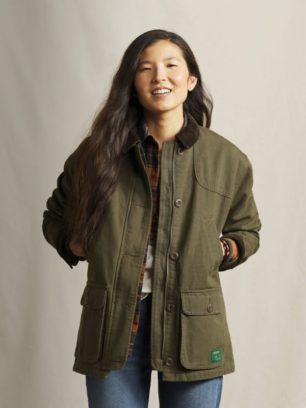 A model wearing an olive green jacket over a plaid shirt and blue jeans.