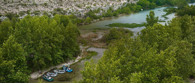 An aerial view of Devils River, winding through lush green trees with rocky hills in the riverbanks with a group of anglers and inflatable rafts pulled up on a gravel bar.