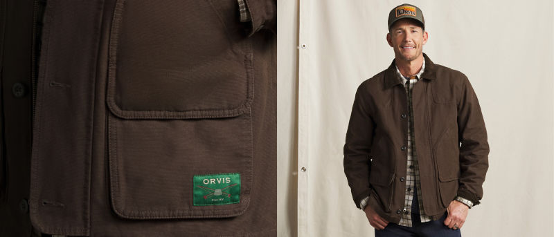 A male model with an Orvis baseball cap wearing a brown jacket over a plaid shirt and dark jeans.