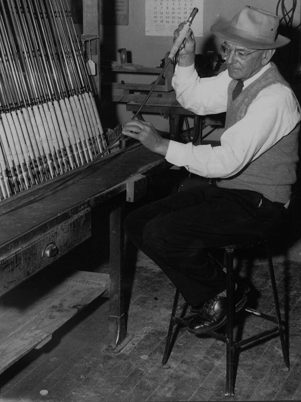 A historical black and white photo of a man in old-fashioned clothing working on a fly rod next to a rack of many rods.