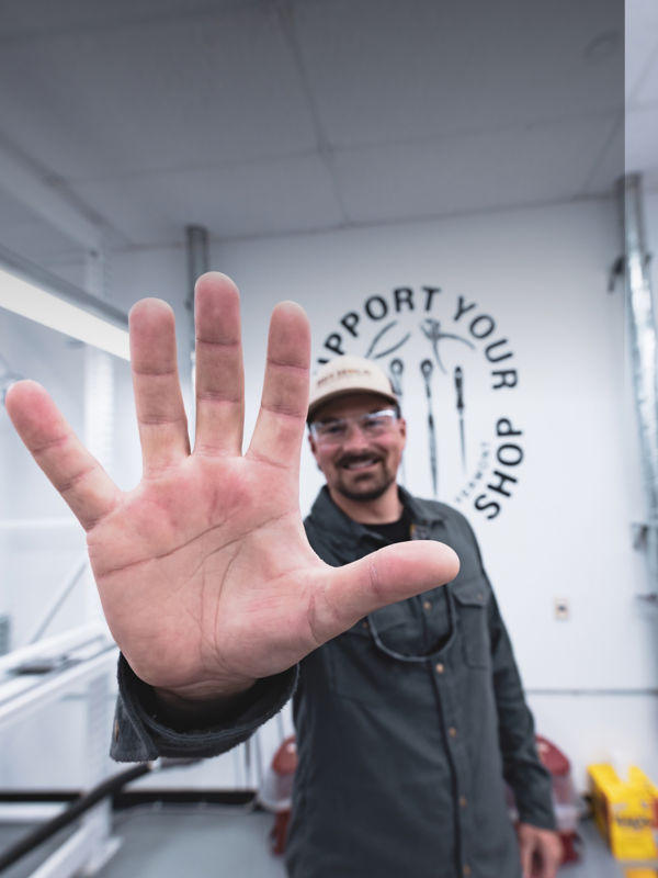 A hand front and center showing 5 fingers with a smiling Orvis Rod Shop employee standing in the rod shop.