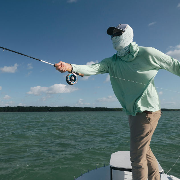 An angler in a sun protective shirt and buff leans into the cast from the front of a boat.