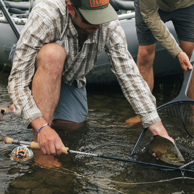 An angler kneels in a stream to release a fish from another angler's net.