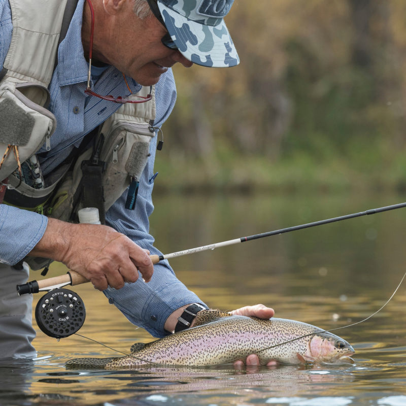 An angler holding a fish in one hand and fly rod in another bends down to release the fish.