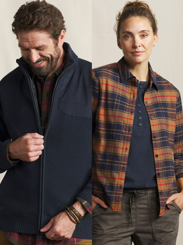 A closeup of a male model wearing a navy blue zip up jacket and a female model wearing a plaid shirt over a navy blue henley.