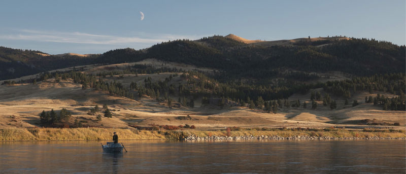 An angler stands in a small boat, surrounded by rolling forested hills and mountains.