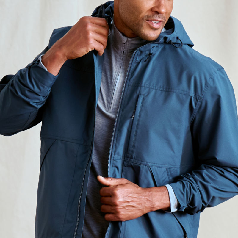 A model stands against a canvas drop cloth wearing a blue rain jacket over a quarter-zip tech shirt.