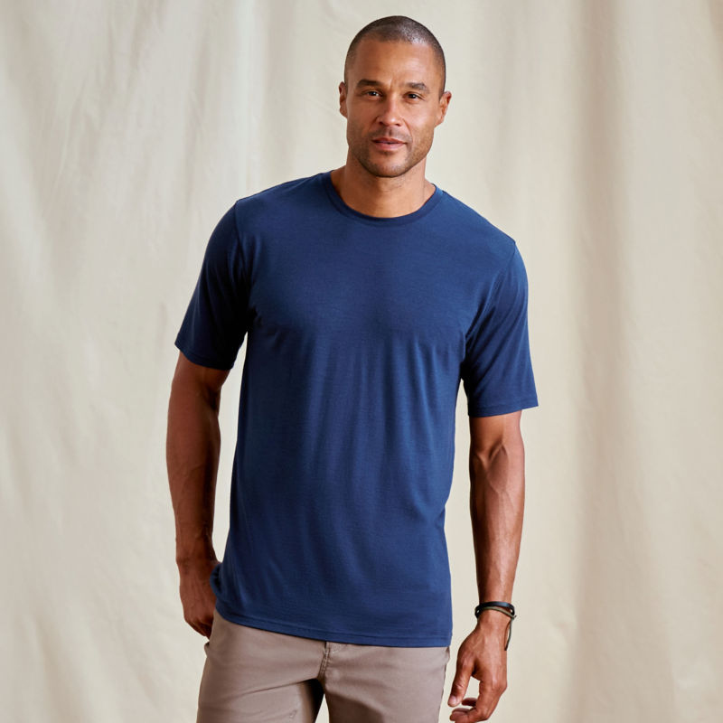 A model stands against a canvas drop cloth wearing a blue merino t-shirt.