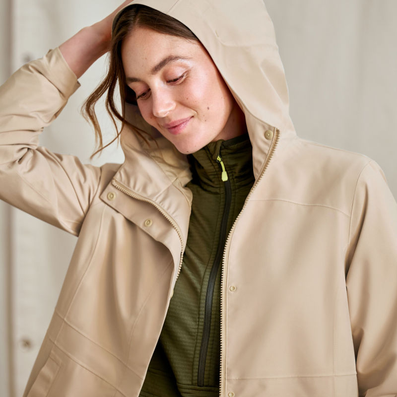 A model stands in front of a canvas drop cloth wearing a cream rain jacket with the hood upright.
