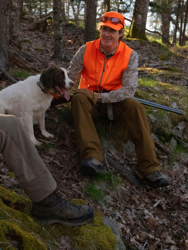 Reid Bryant, Orvis Adventures Endorsed Operations Manager & host of The Orvis Hunting and Shooting Podcast sitting with a dog in the woods on a hunt. the image includes a quote: &ldquo;The best bird hunts are made up of miles, tough terrain, and thorny cover. The Softshell collection goes the distance, keeping me comfortable and protected the whole way.&rdquo;
