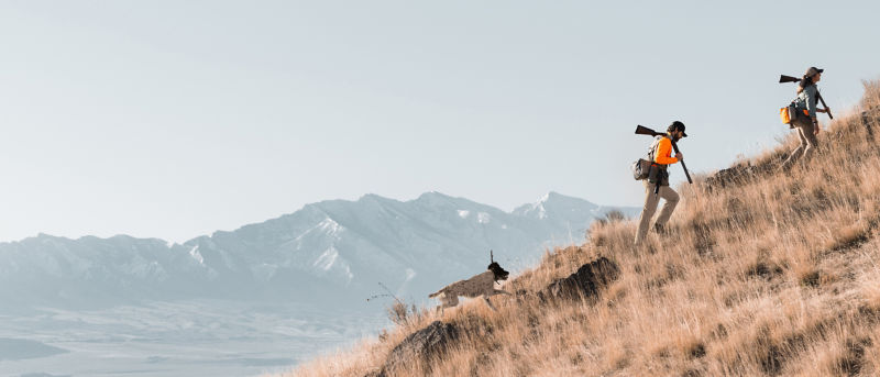 Two hunters hiking up a dry grassy hillside carrying rifles with a mountain range in the distance.