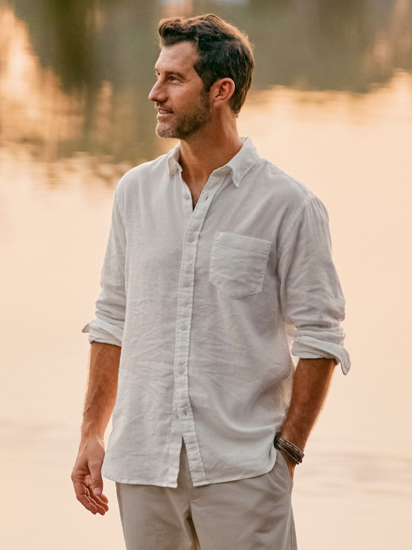 A model stands in front of still water wearing a white linen shirt and khaki shorts.