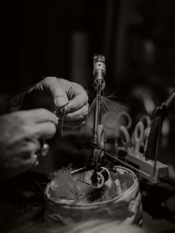 In black and white, hands work to tie a fly on a vise.