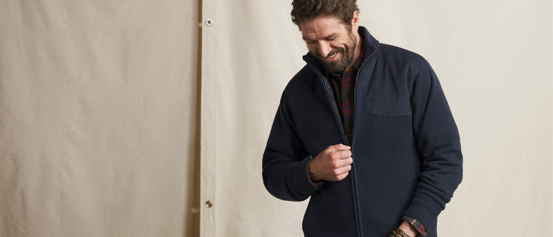 A model standing against an off-white textured background zipping up a dark blue sweater.