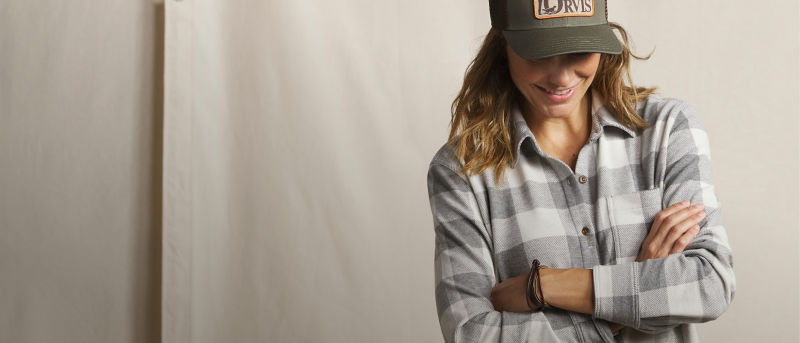 A model with arms her crossed standing against an off-white textured background wearing a gray plaid shirt and an Orvis baseball cap.