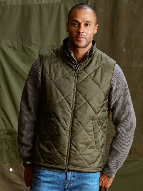 A model wearing an olive quilted Barbour vest over a gray sweater.