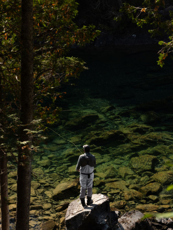 An angler in waders fishes from a huge rock overlooking clear emerald waters.