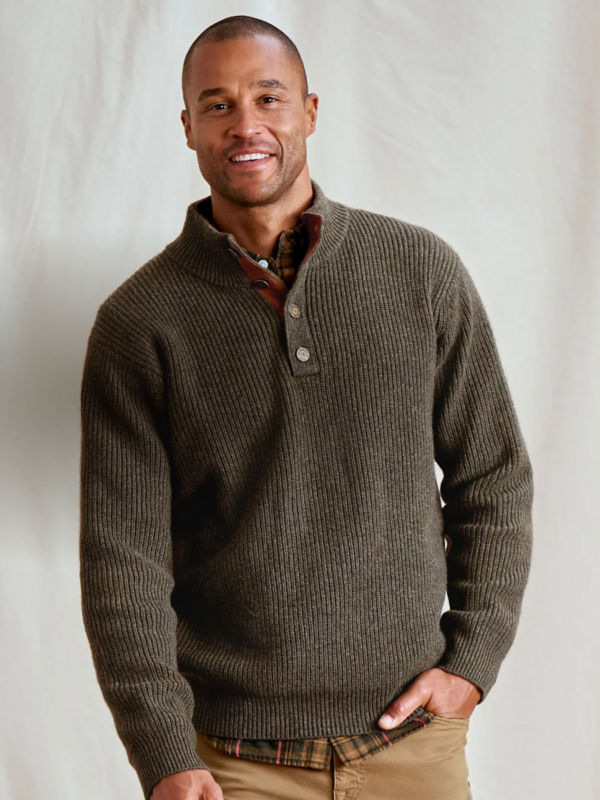 A smiling man stands with one hand in his pocket while wearing a mushroom gray sweater.