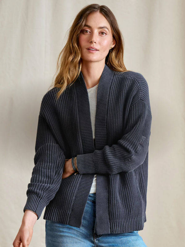 A woman wearing a navy cardigan over a white tee stands in front of a backdrop.