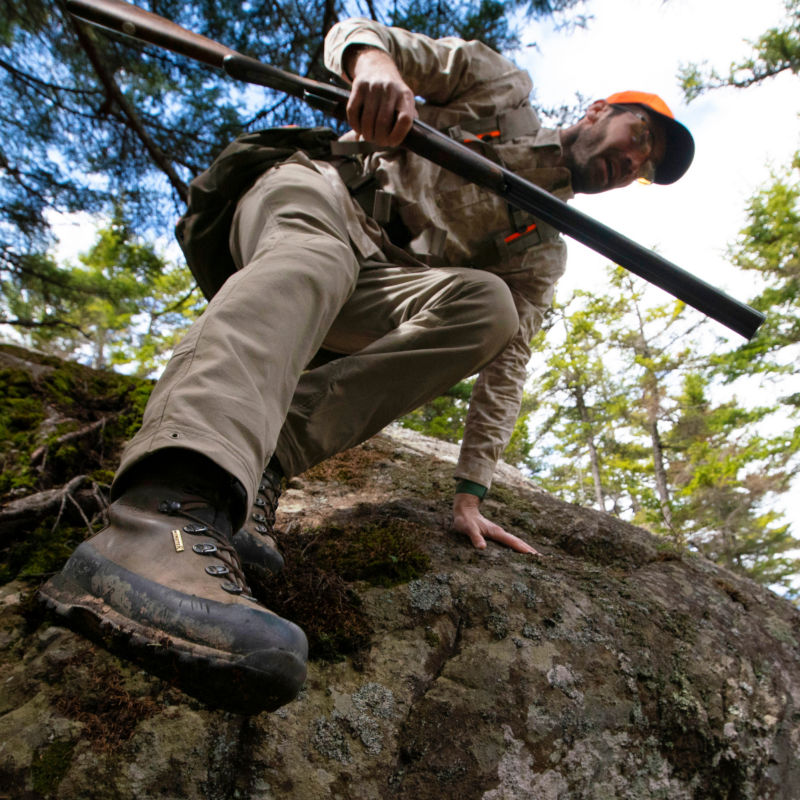 A view from below of a hunter climbing down an embankment holding his shotgun.