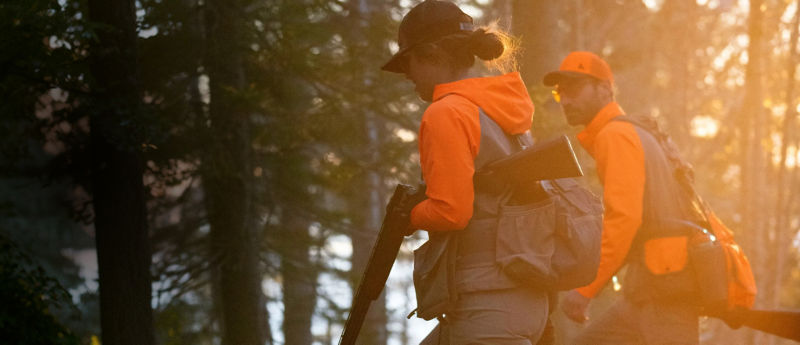 Two hunters in jackets with blaze orange walking through the woods carrying rifles.