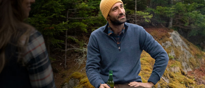 Charley Perkins sits beside a metal fire pit, holding a green bottle while talking with a woman in the foreground.