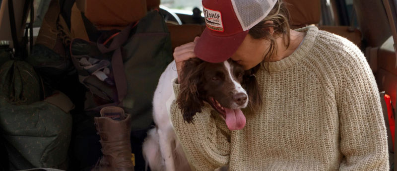 A person wearing a maroon Orvis baseball cap and an oatmeal sweater giving her brown and white spaniel a kiss on the head.