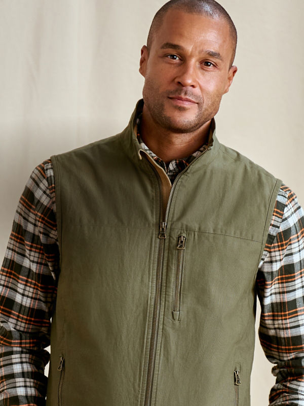 A man  wearing an olive vest over a multi-colored plaid shirt.