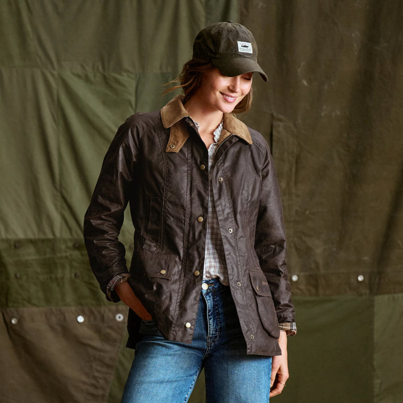 A smiling woman wears a waxed cotton Barbour jacket and ball cap.