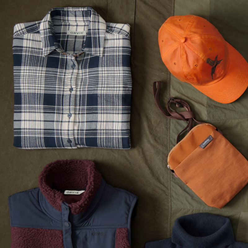A collection of Orvis gifts for women laid neatly on a green background including a blaze orange cap and a crossbody bag.