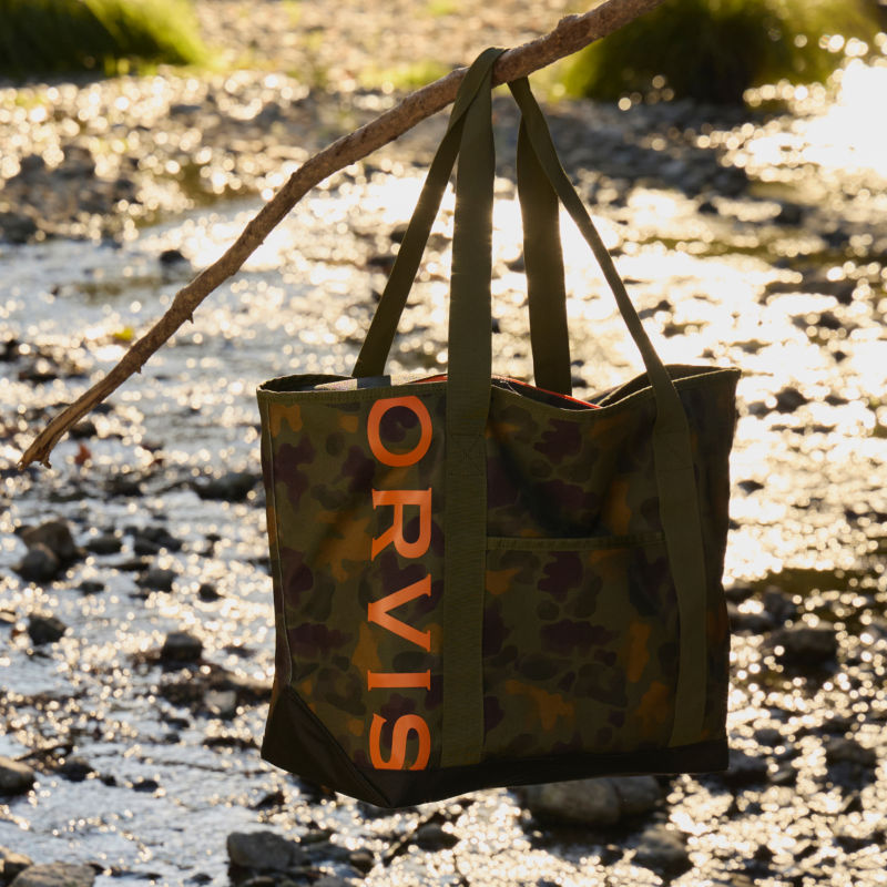 A camo tote bag hanging from a tree by a stream.