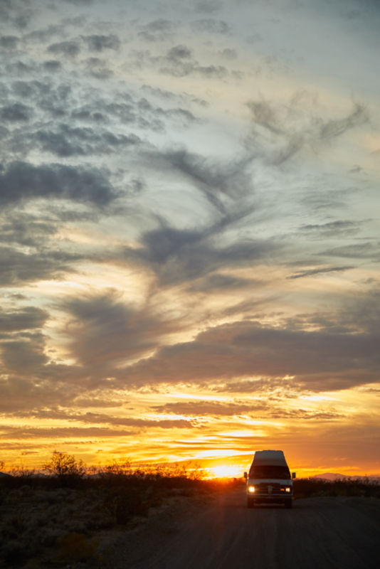 A van with cow skull on hood driving away from setting sun in deseert