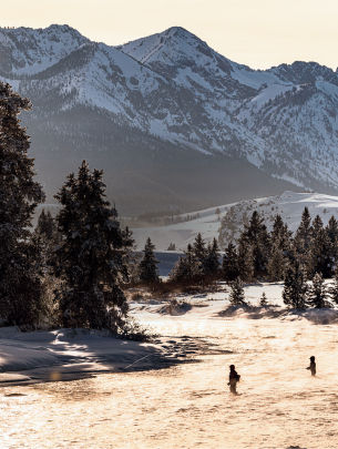 Two anglers fishing in a river with snowy banks and mountains all around.