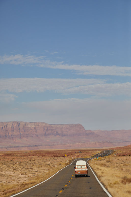 A van trundles down the highway under a giant blue sky, buttes in the background.