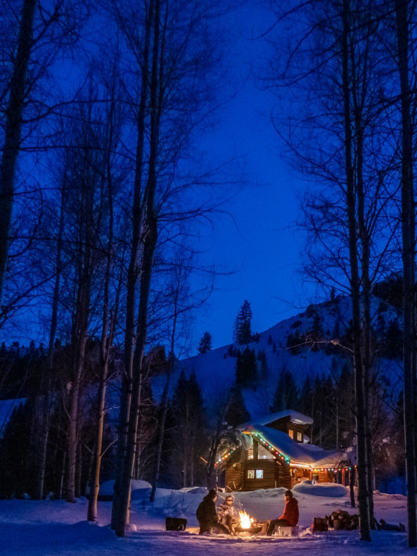Three people sitting around a campfire near a log cabin in the snow.